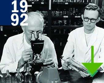 1992. A black and white picture of two men in white lab coats working on a table. One looking into a microscope and the other man is holding a bottle in his hands.
