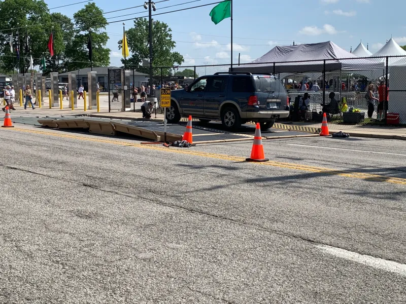 A car driving over a lowered DETER device near a fenced area with tents in it. The device is approximately two car lengths long and one foot above the ground, on a street in front of a chain link fence. There are a few pedestrians about and four traffic cones surrounding the device.