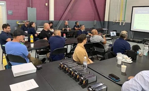 14 people are seated at large folding tables in a large, cinderblock space. There is a row of 8 batteries on one of the tables. They are looking at a presentation projected on a screen.