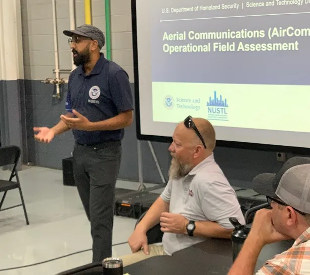 A person stands in front of a screen with the words, “Aerial Communications (AirCom Operational Field Assessment”, and below that, “Science and Technology, NUSTL logos”. There are two other people seated to the speaker’s left.