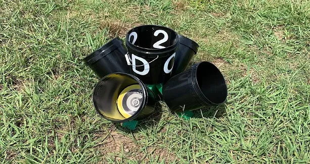 A cluster of medium-sized black plastic buckets sits in a grassy field. There is a circular design visible in one of the buckets and the number 2 in an adjacent bucket, which also has the letters D and O on the outside.