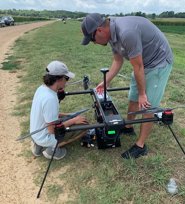 A large drone is resting on a grassy roadside. One person is squatting on the left looking at it, one person is standing bent over on the right gesturing at it.