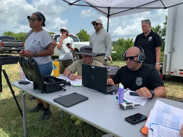 Two people are seated outside in a field at a folding table looking at computer equipment screens. The one on the right is wearing a headset. To the right of the table a person is standing holding a game controller. Behind them three other people are standing facing forward and looking over their heads into the distance.