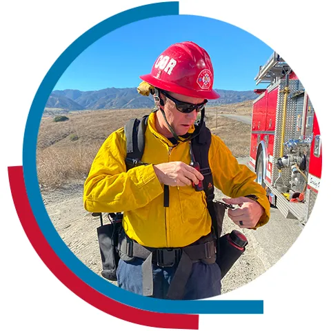 A first responder in full gear standing next to a fire truck out in the field.