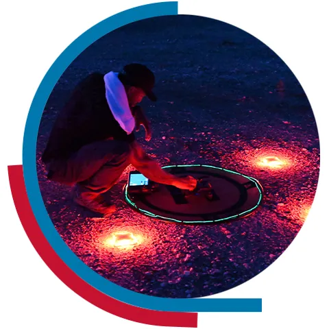 A man setting up a drone on the ground at night marking a circle around the drone with several lights around it.