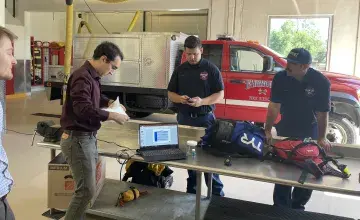 A TDA staff member (wearing a purple collared shirt and handling paperwork) shows two Fairmount firefighters and emergency medical responders (wearing black polo shirts and standing in front of a Fire Rescue vehicle) how to use the wearable chemical sensor badge.