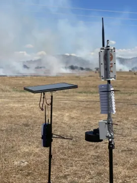 A picture of a flat device on a thin support on the left and three small devices on a thin support next to it on the right. They are standing in a field of burned grass with smoke in the distance.Two fire sensor prototypes during a field test.